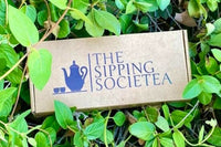 The Sipping Societea Loose leaf tea subscription logo box set in a green leafy garden ready to open up.