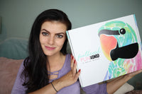 A woman holding up a closed Feathered Fun subscription box.
