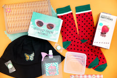 A picnic themed box on a yellow background with watermelon socks, ant picks, a juice pouch, butterfly hat, and bag.