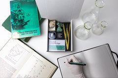 An open Matter subscription box filled with scientific items and tools, next to a person writing in a journal.
