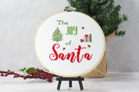 the joy of santa modern cross stitch design with christmas tree and presents on embroidery fabric inside wood hoop