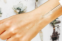 Photo of the back of a hand and a clear glass dropper applying the Buddy System Face Serum by Greenmade goods.