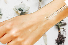 Photo of the back of a hand and a clear glass dropper applying the Buddy System Face Serum by Greenmade goods.