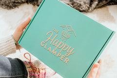 A woman holding a mint green subscription box labeled The Happy Glamper.