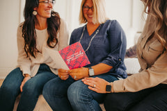 two adult women smiling and looking at another adult women receiving a happy birthday greeting card 