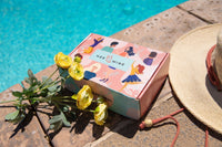 A HER-MINE box placed invitingly next to a sparkling crystal blue swimming pool, surrounded by a vibrant cluster of yellow flowers.