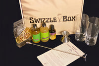 The Queen's Park Swizzle cocktail kit is showcased next to the Swizzle Box box. Two glasses, syrup, bitters, and metal swizzle stick.