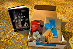 Book titled "The Book Eaters" by Sunyi Dean stands next to open box filled with wrapped book and goodies, laying in orange leaves