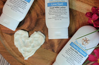 Picture showing three bottles of goat milk lotion on a wooden board with pink flowers. A heart is in the middle drawn with the lotion