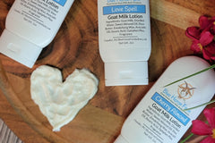 Picture showing three bottles of goat milk lotion on a wooden board with pink flowers. A heart is in the middle drawn with the lotion