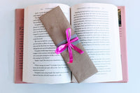 A bookmark wrapped in brown craft paper and decorated with ribbons rests on an open hardcover book. The bookmark is handmade and painted.