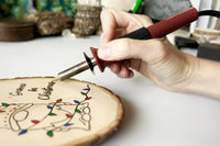 Beginner woodburning tool being held as it burns a Christmas Crate Club project - a gnome wrapped in Christmas lights.