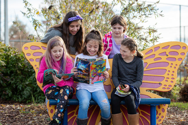 Five girls sit outside, wearing brightly colored clothing. They are on a bench shaped like a butterfly and are reading comic books