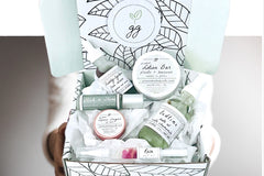 Hands holding a Greenmade goods Beauty Box. Box includes a face serum, lip tin, nail pen, essential oil roller, bath oil, lotion bar.