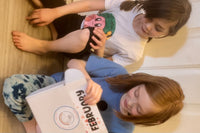 Two girls sit side-by-side and eagerly open their box (marked "FEBRUARY.") One is smiling, one looks curious.