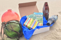 A beach scene with a subscription box from BookTIDE featuring great reads and colorful glass floats