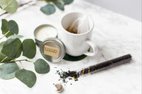 An open tin candle sits beside a tea mug and a vial of open tea. Some of the tea has spilled from the vial.
