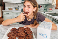 Smiling girl enjoying freshly baked chocolate cookies made with a BāKIT Box baking kit in a home kitchen.