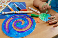 A child's hand colors a circular piece from the Little Dreamers Club Crafts Subscription Box with markers and craft materials
