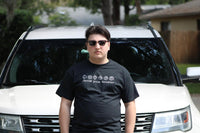 Young man in front of a white SUV. He is wearing sunglasses and a Dungeons and Dragons shirt with dice and the words "Choose your Weapon".