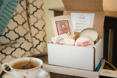 A box holding a small candle, pouch of tea, and matches sits beside a cup of tea on a side table. A cozy chair is in the background.