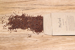 A brown paper Sproutie seed packet lies on a wooden background. Mustard sprouting seeds lay across the table.