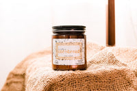 a candle named as country as cornbread is sitting on an empty burlap bag that is spread across a table.