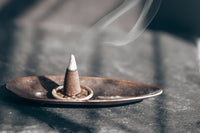 grey platform with oblong brown bowl. inside the bowl is a white round disk with a brown cone incense that is lit and burning