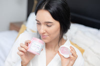 brunette woman in a white bath robe smelling an open whipped soap scrub while sitting on a white bed
