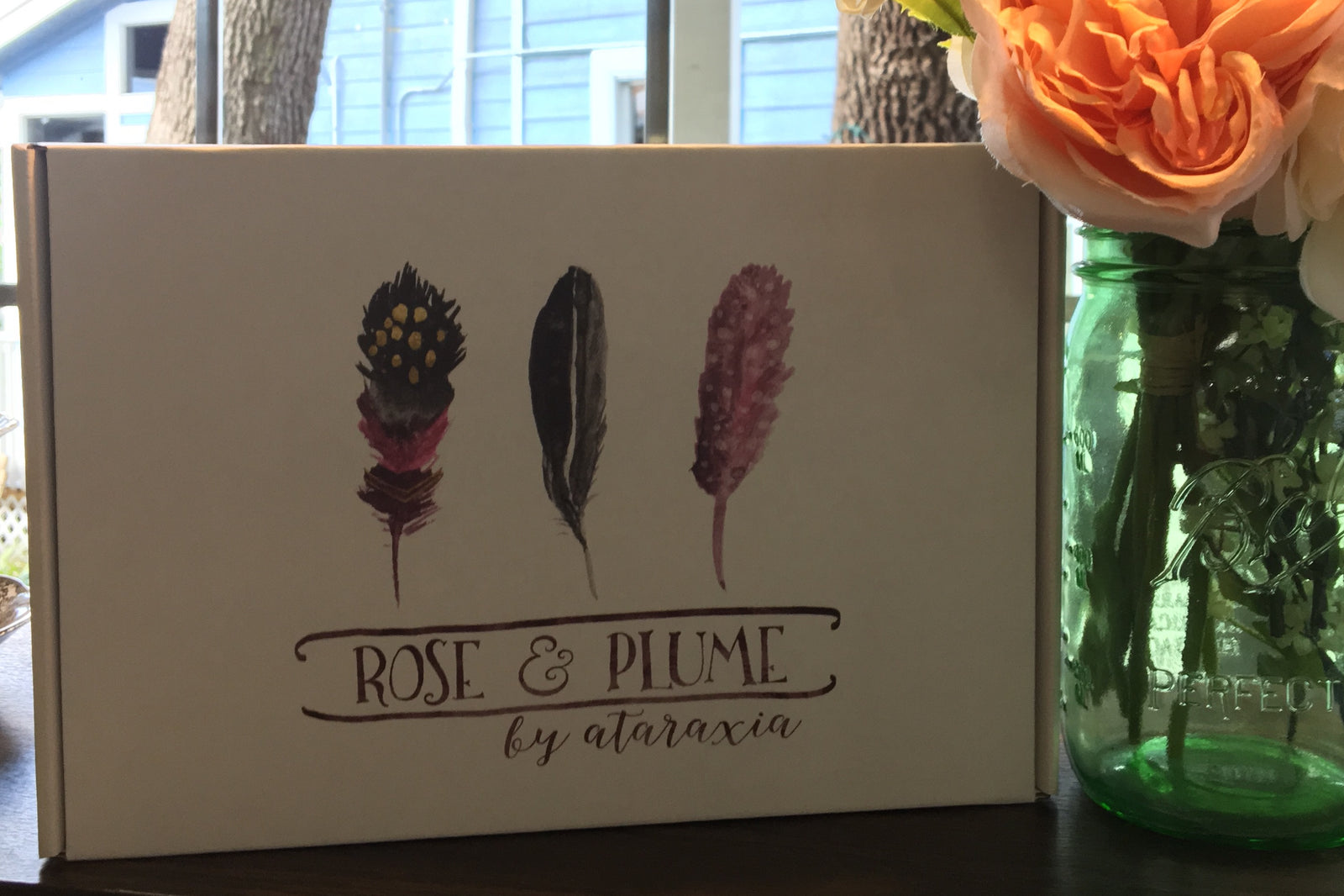 A closed Rose and Plume subscription box next to a mason jar full of pink flowers.