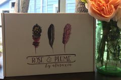 A closed Rose and Plume subscription box next to a mason jar full of pink flowers.