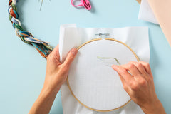 lady stitching white counted cross stitch fabric in size 14 count mounted inside wood embroidery hoop on green background