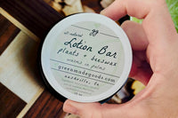 Hand holding a Sandalwood Lotion Bar tin by Greenmade goods with wood slats and plants in the background.
