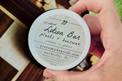 Hand holding a Sandalwood Lotion Bar tin by Greenmade goods with wood slats and plants in the background.