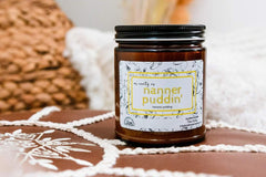 a jarred candle named nanner pudding sits on a brocade stool. a wicker basket and some off white material is in the background.