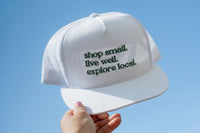 A hand holding a white cap with green text saying "shop small, live well, explore local" for the Explore Local Box monthly subscription