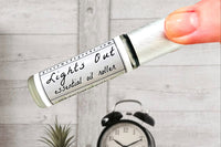 Two fingers holding the Lights Out Essential Oil roller bottle with an alarm clock on a bedside table in the background.