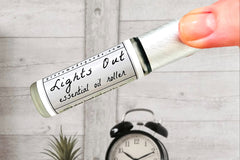 Two fingers holding the Lights Out Essential Oil roller bottle with an alarm clock on a bedside table in the background.