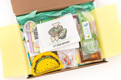 A taco-themed subscription box open. Items inside taco coin purse, avocado greeting card, dip packet, colorful earrings, and pitter tool.