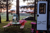 That Road Trip Mom Escape and Explore Box displayed on a picnic table at a RV campsite at with trees and a sunset in the background
. 

