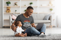 A young dad with his little baby sitting on a rug on his laptop. He is wearing a Star Wars Fighter Tshirt.