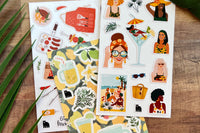 A selection of summer themed stickers including BBQ, lemonade and women, shown on a wooden desk with fronds overlapping.  