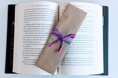 A bookmark wrapped in brown craft paper and decorated with ribbons rests on an open hardcover book. The bookmark is handmade and painted.
