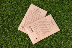 Two seed packets sit on top of each other with a grass background. They are brown paper. One says "kale" and the other "corn".