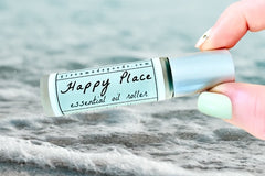 Two fingers holding the Happy Place Essential Oil roller bottle with foamy ocean waves in the background.