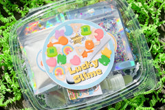 Lucky Slime groovy slime kit from Bored to Brilliant in a clear deli container with glitter and clay pieces, slime activator, jello powder.