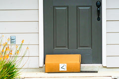 Front doorstep is pictured. Door is black with a black handle. On the doorstep rests a cardboard box with Attempting Less logo imprinted.