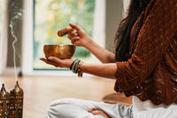 woman sitting cross legged with a sound bowl and paddle holding it up over a brass incense burner with smoke wafting up