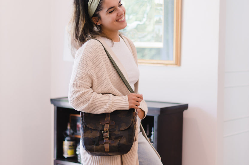 A smiling woman wearing a cream knit cardigan and a green headband carries a dark camouflage messenger bag over her shoulder.