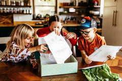 three children are sitting at a dining room table. they are opening up a wiggle worms kit box. they are laughing.